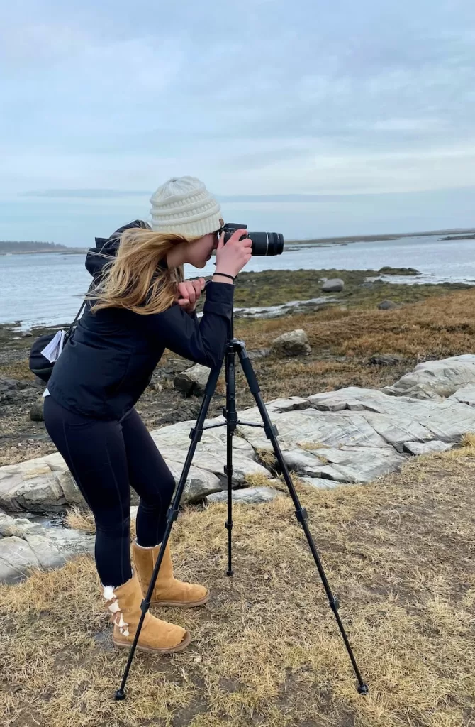 woman in black jacket and blue denim jeans sitting on rock near sea during daytime with a lightweight backpacking tripod